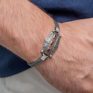 Peut inclure: Bracelet en argent avec une pièce maîtresse rectangulaire ornée de trois pierres rouges. La pièce maîtresse présente des accents dorés et des détails ornés. Le bracelet est porté sur un poignet sur fond bleu.