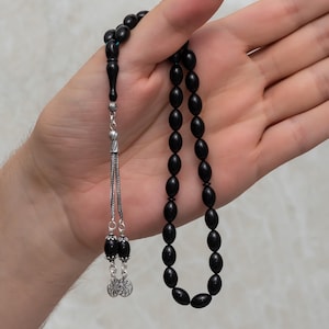 Peut inclure: Chapelet noir avec des perles ovales. Les perles sont enfilées sur une chaîne argentée et des éléments décoratifs. Le pompon comporte deux perles noires et deux breloques argentées en forme de fleur.