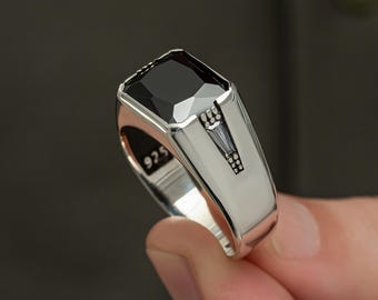 May include: A silver ring with a large, rectangular black gemstone. The ring has a polished finish and features a decorative design on the band. The ring is marked with "925" on the inside.