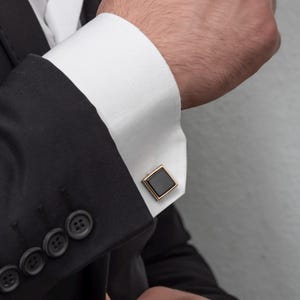 May include: Close-up of a black suit with a white dress shirt and gold-toned square cufflinks. The cufflinks have a black square center and a gold-colored border. The suit has black buttons.