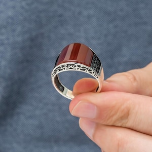 May include: A silver ring featuring a rectangular, reddish-brown stone. The ring has a decorative, patterned band and a black stone accent. The ring is held against a blue background.