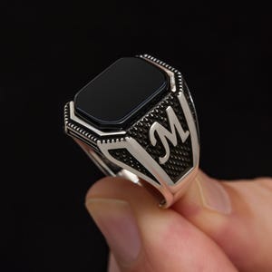 Könnte beinhalten: Ein silberner Ring mit einem rechteckigen schwarzen Onyxstein. Der Ring hat ein detailliertes Design am Band, einschließlich des Buchstabens "M". Der Ring wird vor einem schwarzen Hintergrund gehalten.
