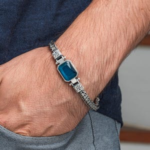 May include: Silver bracelet featuring a rectangular, teal-colored gemstone. The bracelet has a woven chain design with decorative accents near the gemstone. The jewelry is displayed on a person's wrist, showcasing the bracelet's details.