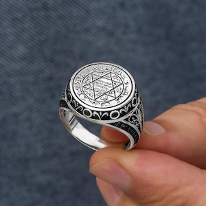 May include: A silver ring with an engraved design. The ring features a central star within a circle, surrounded by intricate patterns and text. The band has a detailed, ornate design. The ring is held against a grey background.