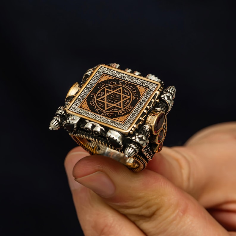 May include: A silver ring with a square, ornate face featuring a gold and black design with a star symbol. The ring has intricate detailing and small red accents. The ring is held up by a person's fingers.