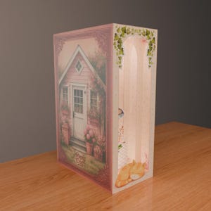 Puede incluir: Caja decorativa de papel rosa con una escena de caba&ntilde;a, puerta blanca y detalles florales. El lateral presenta un dise&ntilde;o arqueado y una ilustraci&oacute;n de gato naranja durmiendo. El texto "Paply Craft" es visible.