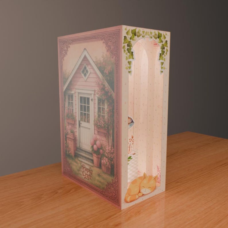 Puede incluir: Caja decorativa de papel rosa con una escena de caba&ntilde;a, puerta blanca y detalles florales. El lateral presenta un dise&ntilde;o arqueado y una ilustraci&oacute;n de gato naranja durmiendo. El texto "Paply Craft" es visible.