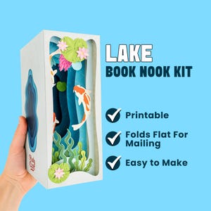 Könnte beinhalten: Ein weißes Lake Book Nook Kit mit einer Papierbastel-Seelandschaft mit Koi-Karpfen, Seerosen und Wasserpflanzen. Der Text "LAKE BOOK NOOK KIT" ist in Weiß dargestellt. Das Kit ist mit "Druckbar", "Flach zum Versenden falten" und "Einfach zu machen" gekennzeichnet.