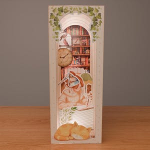 Puede incluir: Una caja decorativa iluminada con un dise&ntilde;o en capas que representa una escena de lectura. La caja incluye una estanter&iacute;a, una persona escribiendo, un gato y un reloj. La caja rectangular tiene un marco blanco con detalles de hiedra verde.