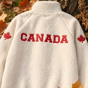 Peut inclure: Veste polaire crème avec un col montant. Le mot "CANADA" est brodé en rouge dans le dos, flanqué d'emblèmes de feuilles d'érable rouges. La veste a un aspect doux et texturé.