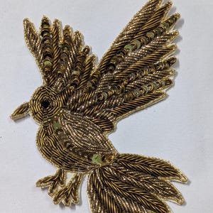 May include: A bird-shaped patch featuring intricate gold metallic thread embroidery and small, reflective gold sequins. The design depicts a bird in flight with wings spread, detailed feather textures, and a dark bead eye on a white fabric backing.