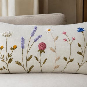 Può includere: Un cuscino rettangolare color bianco sporco con ricami dettagliati di vari fiori di campo. Il design presenta steli floreali gialli, viola, rosa e blu con foglie verdi e petali bianchi sulla superficie anteriore.