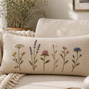 May include: A rectangular beige throw pillow featuring detailed embroidery of various wildflowers including lavender, daisies, and cornflowers. The stems and leaves are stitched in green thread, while the flower heads display shades of yellow, purple, pink, white, and blue.
