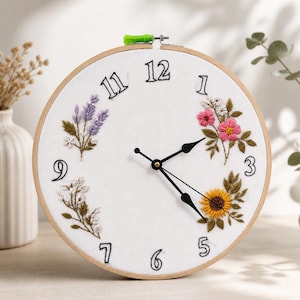 Op de afbeelding: Een ronde klok met een witte wijzerplaat, zwarte cijfers en wijzers. De wijzerplaat is versierd met geborduurde bloemen in roze, geel en paars. De klok is geplaatst in een houten ring.