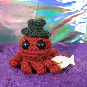May include: A handmade red crocheted octopus wearing a small gray top hat. The octopus has large black eyes and is sitting on a purple surface. A tag in the shape of a fish is attached to the octopus.