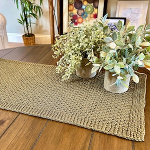 May include: A rectangular, olive green knitted table runner. The runner is placed on a round, wooden table. Decorative plants in small, concrete pots are arranged on the table. The background includes a sofa and framed artwork.