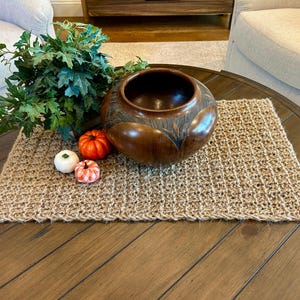 May include: A dark brown wooden bowl with a leaf design sits on a woven jute placemat. Miniature pumpkins in orange, white, and orange-and-white patterns and green foliage complete the autumn-themed arrangement on a wooden table.