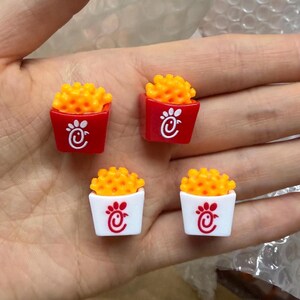 May include: Four miniature resin charms shaped like containers of waffle fries. Two containers are red and two are white, each featuring a small white or red chicken logo on the front. The fries are textured, bright yellow, and rounded in shape.