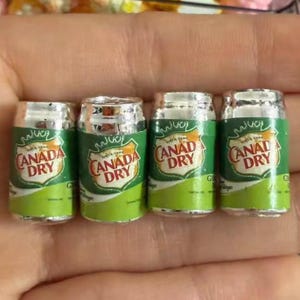 May include: Four miniature beverage cans featuring the Canada Dry logo. Each small, cylindrical container is coloured in shades of green, white, and gold, with the text Canada Dry clearly printed on the front of the shield-shaped emblem.