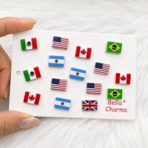 May include: A collection of small rectangular flag charms arranged on a white display card. The set includes the flags of Mexico, the United States, Canada, Brazil, Argentina, and the United Kingdom. The text Bella Charms is printed in pink.