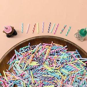 Puede incluir: Un cuenco de madera grande lleno de una multitud de palitos finos y pequeños con rayas multicolores. Dos pequeñas figuras con forma de oso, una con un sombrero rosa y otra con un sombrero verde, flanquean el cuenco, con diez palitos alineados.