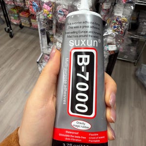 May include: A grey tube of B-7000 multi-purpose adhesive with a white nozzle. The label features the text suxun, B-7000, Gravity magic rubber, and 3.7fl.oz(110mL). The red bottom section lists features including waterproof, flexible, and anti-vibration.