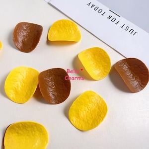 May include: A collection of small, curved, textured charms shaped like potato chips in yellow and brown colors. The charms are scattered across the surface, with some pieces featuring a matte finish. The text Bella Charms is printed in pink.