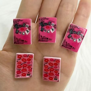 May include: Five rectangular flat charms featuring a bright pink background. Three charms display the text BURN BOOK in a collage style with black letters on white squares. Two charms feature a repeating pattern of red lip prints and black scribbles.