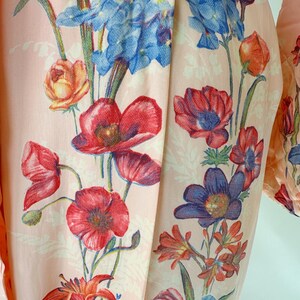 May include: Close-up of a light pink fabric with a floral pattern. The design features red poppies, blue irises, and orange lilies. The flowers are set against a backdrop of smaller, white floral motifs.