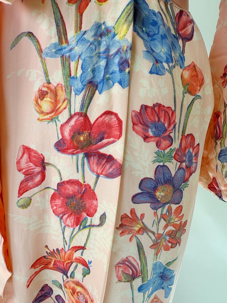 May include: Close-up of a light pink fabric with a floral pattern. The design features red poppies, blue irises, and orange lilies. The flowers are set against a backdrop of smaller, white floral motifs.