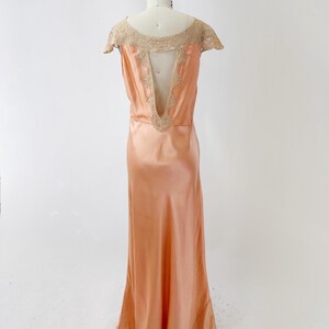 May include: Peach-colored vintage silk gown on a mannequin. The dress features a low back with lace trim and cap sleeves. The gown has a flowing skirt and lace detailing at the hem. The dress is a classic, elegant design.