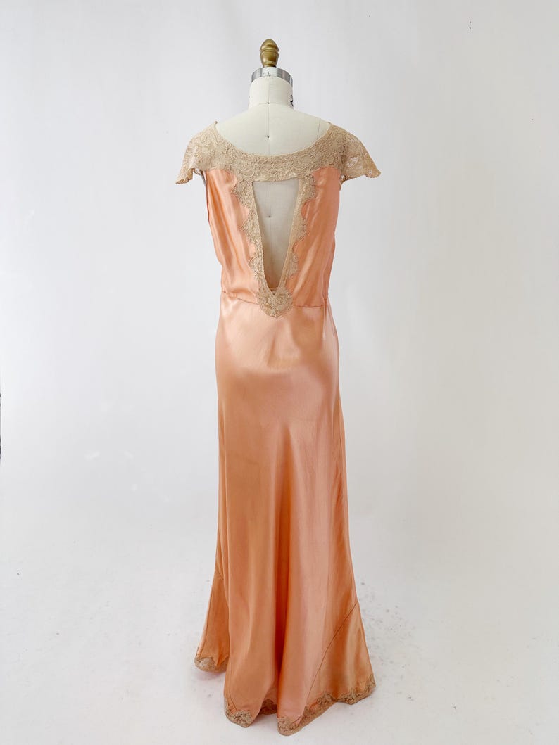 May include: Peach-colored vintage silk gown on a mannequin. The dress features a low back with lace trim and cap sleeves. The gown has a flowing skirt and lace detailing at the hem. The dress is a classic, elegant design.
