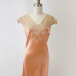 May include: A peach-colored vintage slip dress with a V-neckline and delicate lace detailing. The dress features a flowing silhouette and intricate embroidery on the bodice, showcasing a classic, elegant design.