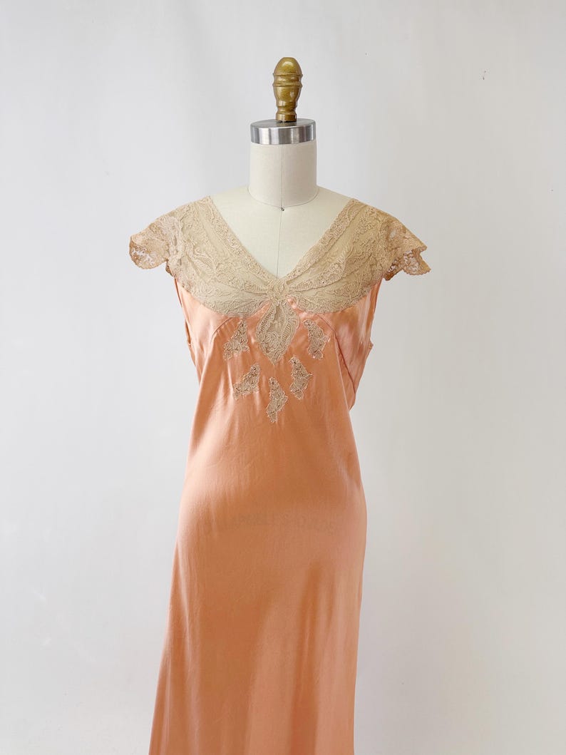 May include: A peach-colored vintage slip dress with a V-neckline and delicate lace detailing. The dress features a flowing silhouette and intricate embroidery on the bodice, showcasing a classic, elegant design.