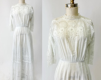 May include: A white vintage dress with intricate eyelet and lace detailing. The dress features a high neckline, three-quarter sleeves, and a tiered skirt. The dress is displayed on a mannequin against a plain white background.