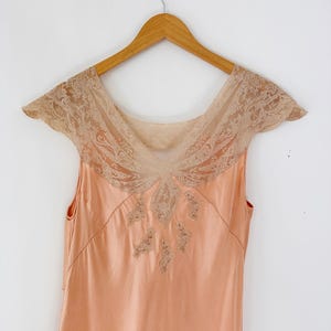 May include: A peach-colored vintage slip dress with a delicate lace neckline and cap sleeves. The dress is sleeveless and hangs on a wooden hanger against a white background. The lace detailing is a light beige color.