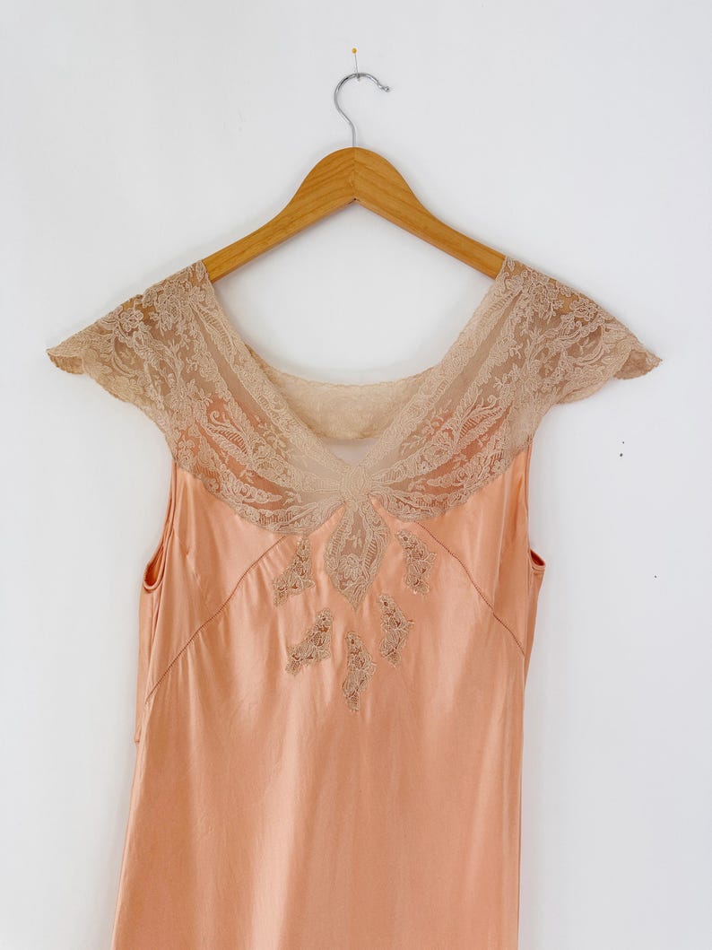 May include: A peach-colored vintage slip dress with a delicate lace neckline and cap sleeves. The dress is sleeveless and hangs on a wooden hanger against a white background. The lace detailing is a light beige color.