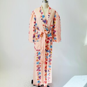 May include: A peach-colored robe with a floral pattern and quilted sleeves. The robe features a wrap-around design with a matching belt. The floral design includes red, blue, purple, and yellow flowers. The robe is displayed on a mannequin.