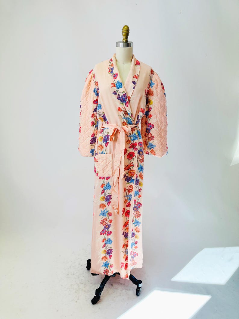 May include: A peach-colored robe with a floral pattern and quilted sleeves. The robe features a wrap-around design with a matching belt. The floral design includes red, blue, purple, and yellow flowers. The robe is displayed on a mannequin.