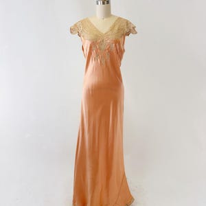May include: Peach-colored vintage slip dress with a V-neckline and delicate lace detailing. The dress features a flowing, floor-length design and is displayed on a mannequin. The lace trim is visible on the neckline and hem.
