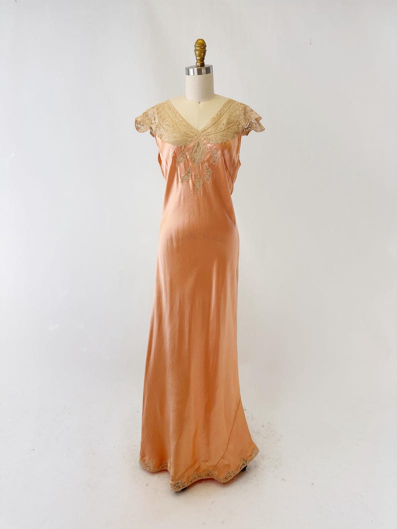 May include: Peach-colored vintage slip dress with a V-neckline and delicate lace detailing. The dress features a flowing, floor-length design and is displayed on a mannequin. The lace trim is visible on the neckline and hem.