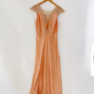 May include: A peach-colored vintage slip dress with a V-neckline and delicate lace detailing on the shoulders and neckline. The dress is hanging on a wooden hanger against a white background. The hemline also features lace.