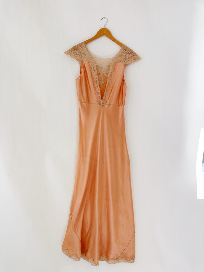May include: A peach-colored vintage slip dress with a V-neckline and delicate lace detailing on the shoulders and neckline. The dress is hanging on a wooden hanger against a white background. The hemline also features lace.