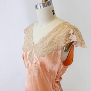 May include: Peach-colored vintage slip dress with a V-neckline and delicate lace detailing. The dress features a sleeveless design and a flowing silhouette. The lace trim is a light beige color, adding a touch of elegance to the garment.