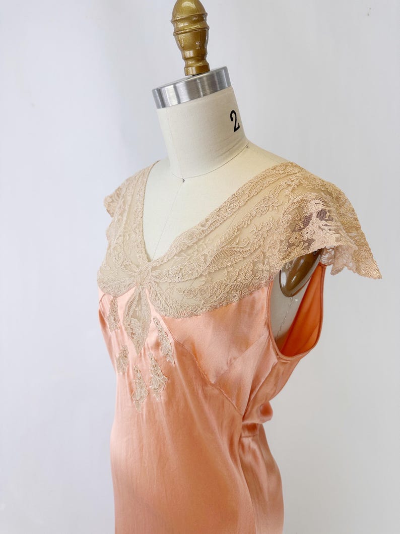 May include: Peach-colored vintage slip dress with a V-neckline and delicate lace detailing. The dress features a sleeveless design and a flowing silhouette. The lace trim is a light beige color, adding a touch of elegance to the garment.