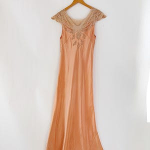 May include: A peach-colored vintage slip dress with a delicate lace neckline and cap sleeves. The dress features intricate embroidery on the bodice and hem, and hangs on a wooden hanger against a white background. The dress has a flowing, floor-length design.