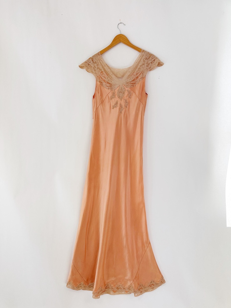 May include: A peach-colored vintage slip dress with a delicate lace neckline and cap sleeves. The dress features intricate embroidery on the bodice and hem, and hangs on a wooden hanger against a white background. The dress has a flowing, floor-length design.