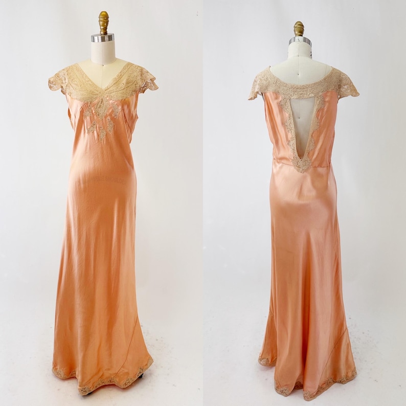 May include: A peach-colored, floor-length vintage dress with a V-neckline and cap sleeves. The dress features delicate lace and embroidery around the neckline and hem. The back of the dress has a low V-shaped opening.