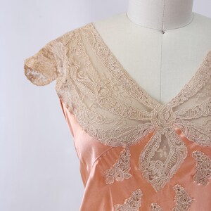 May include: Peach-colored satin slip with intricate beige lace detailing. The slip features a V-neckline and delicate lace accents on the bodice and sleeves. The lace is a contrasting color to the satin, creating a vintage aesthetic.