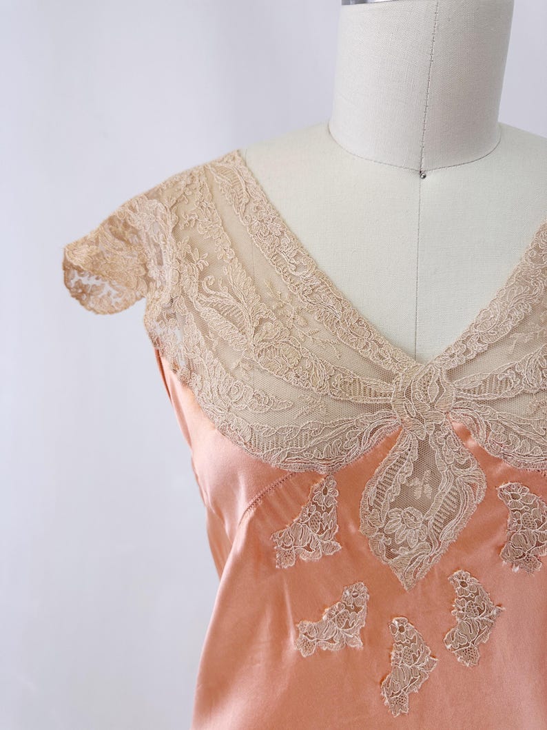 May include: Peach-colored satin slip with intricate beige lace detailing. The slip features a V-neckline and delicate lace accents on the bodice and sleeves. The lace is a contrasting color to the satin, creating a vintage aesthetic.
